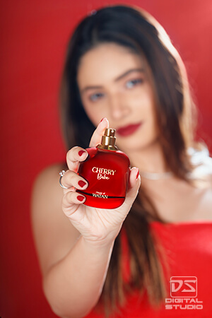 Red perfume bottle held in sharp focus towards the camera, with the model softly blurred against a red background.