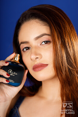Close-up portrait of a model holding a dark blue perfume bottle beside her face against a blue background.